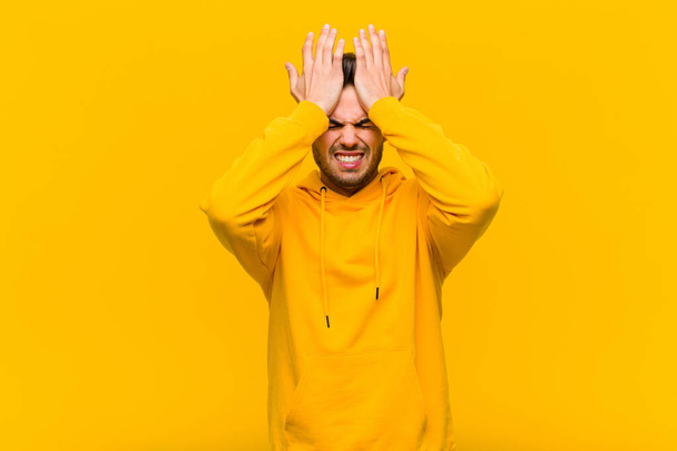 young hispanic man feeling stressed and anxious, depressed and frustrated with a headache, raising both hands to head against orange wall - 写真・画像