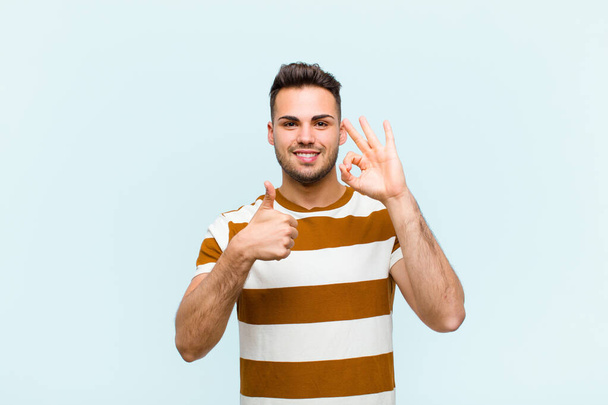 young hispanic man feeling happy, amazed, satisfied and surprised, showing okay and thumbs up gestures, smiling against blue wall - Foto, immagini