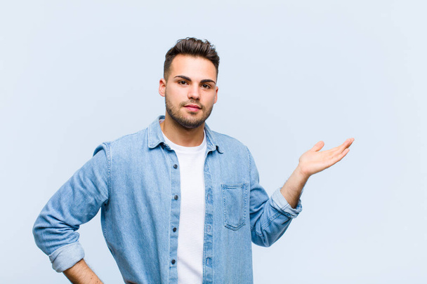 young hispanic man smiling, feeling confident, successful and happy, showing concept or idea on copy space on the side against blue wall - Foto, immagini