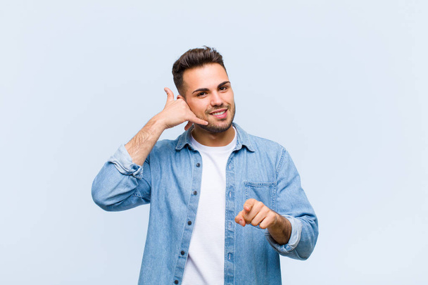 young hispanic man smiling cheerfully and pointing to camera while making a call you later gesture, talking on phone against blue wall - Fotó, kép