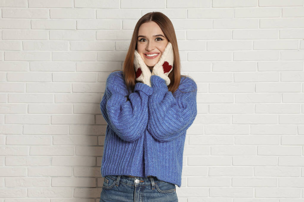Beautiful young woman in mittens and blue sweater near white brick wall. Winter season - Foto, afbeelding