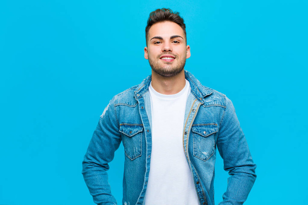 young hispanic man smiling happily with a hand on hip and confident, positive, proud and friendly attitude against blue background - Foto, imagen