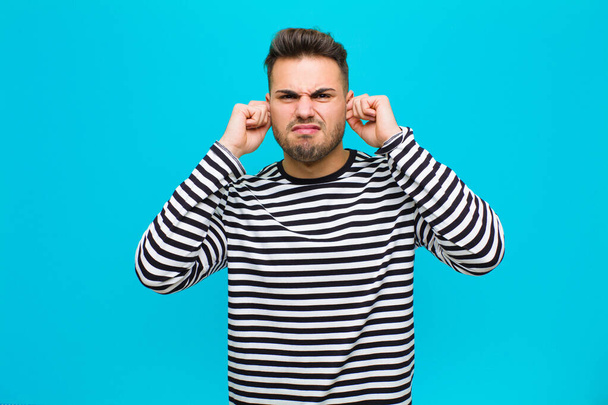 young hispanic man looking angry, stressed and annoyed, covering both ears to a deafening noise, sound or loud music against blue background - Photo, image