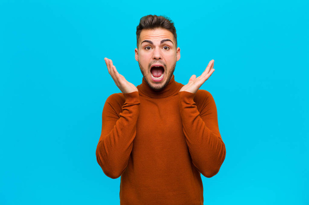 young hispanic man feeling shocked and excited, laughing, amazed and happy because of an unexpected surprise against blue background - Foto, imagen