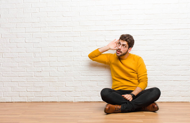 young handsome man sitting on home floor against brick wall texture - Foto, Imagen