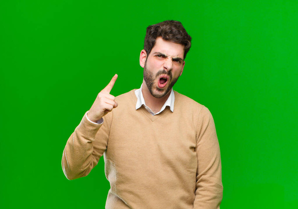 young businessman pointing at camera with an angry aggressive expression looking like a furious, crazy boss against green background - Fotó, kép