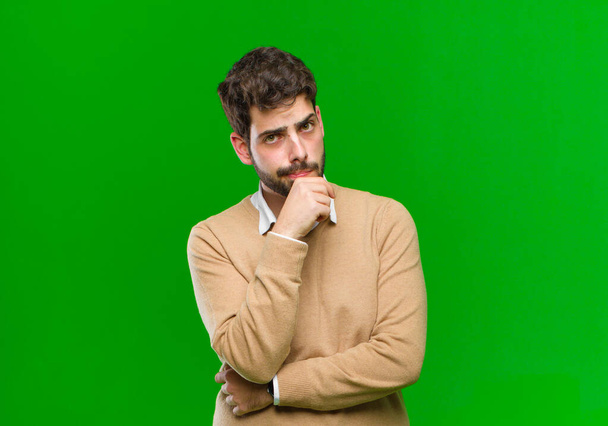 young businessman looking serious, confused, uncertain and thoughtful, doubting among options or choices against green background - Foto, afbeelding