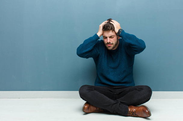 young handsome man sitting on floor feeling stressed and frustrated, raising hands to head, feeling tired, unhappy and with migraine - Photo, Image