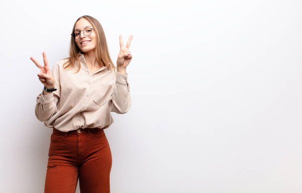 young pretty woman smiling and looking happy, friendly and satisfied, gesturing victory or peace with both hands against white wall - Fotoğraf, Görsel