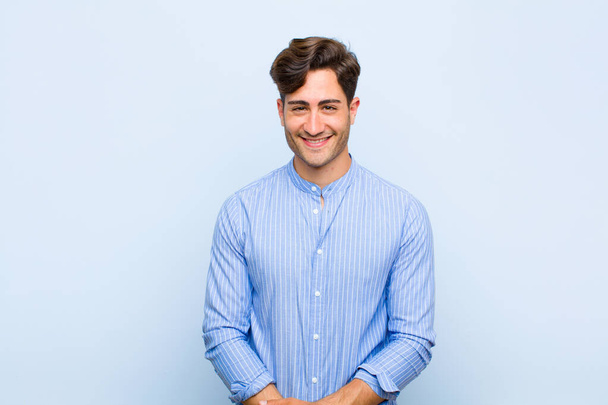 junger gutaussehender Mann lächelt positiv und selbstbewusst, sieht zufrieden, freundlich und glücklich vor blauem Hintergrund aus - Foto, Bild