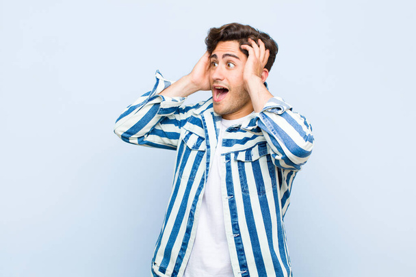 young handsome man feeling happy, excited and surprised, looking to the side with both hands on face against blue background - 写真・画像