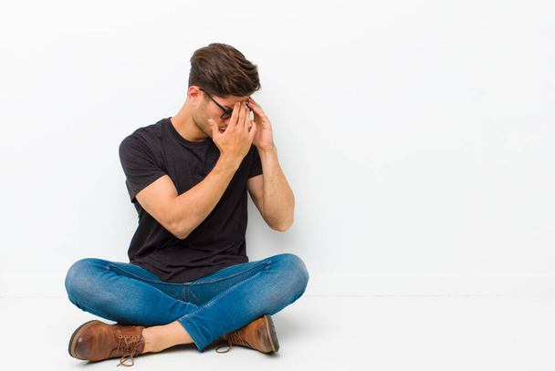 young handsome man covering eyes with hands with a sad, frustrated look of despair, crying, side view sitting on the floor - Photo, Image