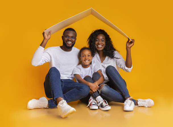 Familia negra sentada bajo techo simbólico soñando con una nueva casa
. - Foto, Imagen