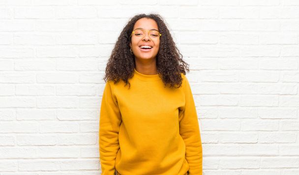 młoda czarna kobieta z dużym, przyjaznym, beztroskim uśmiechem, wyglądająca pozytywnie, zrelaksowana i szczęśliwa, chłodząca się pod ceglaną ścianą - Zdjęcie, obraz