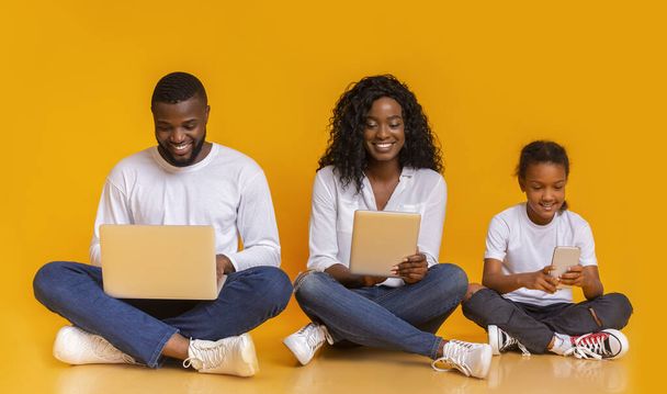 gelukkig zwart familie met behulp van verschillende gadgets, zitten op de vloer samen - Foto, afbeelding