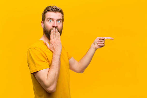 young blonde man feeling happy, shocked and surprised, covering mouth with hand and pointing to lateral copy space against orange wall - Valokuva, kuva