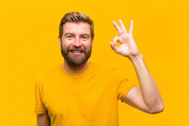 young blonde man feeling happy, relaxed and satisfied, showing approval with okay gesture, smiling against orange wall - Fotó, kép