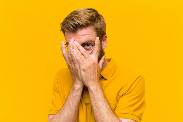 young blonde man feeling scared or embarrassed, peeking or spying with eyes half-covered with hands against orange wall - Photo, image
