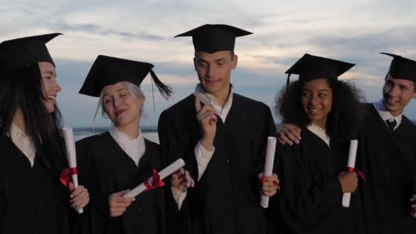 Mixed races happy students in classic graduation mantles and academic hats walking and talking. - Footage, Video