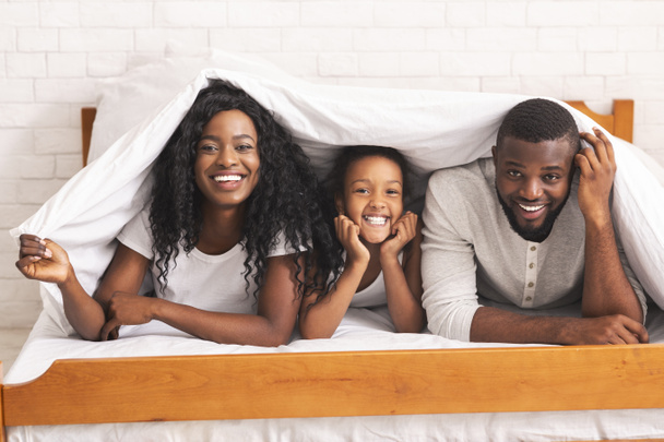 fröhliche schwarze dreiköpfige Familie versteckt sich unter einer Decke auf dem Bett - Foto, Bild