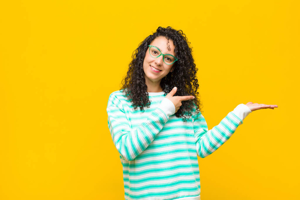 young pretty woman smiling cheerfully and pointing to copy space on palm on the side, showing or advertising an object against orange wall - Foto, imagen