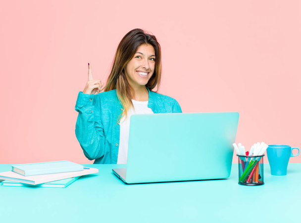young pretty woman working with a laptop smiling cheerfully and happily, pointing upwards with one hand to copy space - 写真・画像
