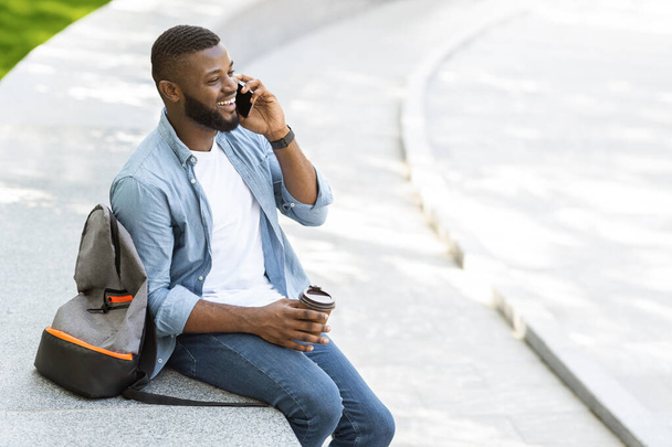 fröhlicher schwarzer Typ, der draußen telefoniert und Kaffee trinkt - Foto, Bild