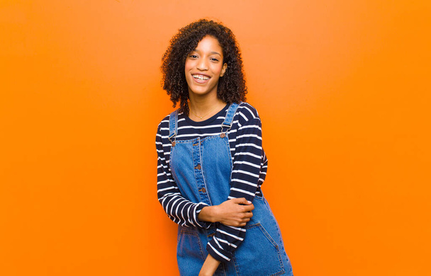 молодая красивая черная женщина смеется застенчиво и весело, с дружелюбным и позитивным, но небезопасным отношением к оранжевой стене
 - Фото, изображение