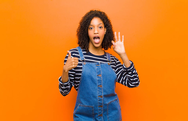 young pretty black woman feeling happy, amazed, satisfied and surprised, showing okay and thumbs up gestures, smiling against orange wall - Fotografie, Obrázek