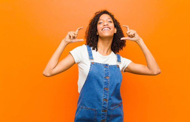 young pretty black woman framing or outlining own smile with both hands, looking positive and happy, wellness concept against orange wall - Фото, изображение