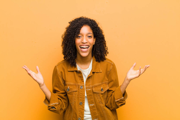 young pretty black woman feeling happy, excited, surprised or shocked, smiling and astonished at something unbelievable against orange wall - Фото, изображение