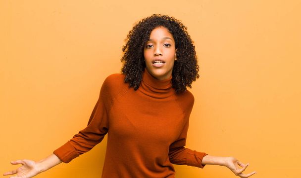 young pretty black woman feeling clueless and confused, having no idea, absolutely puzzled with a dumb or foolish look against orange wall - Zdjęcie, obraz