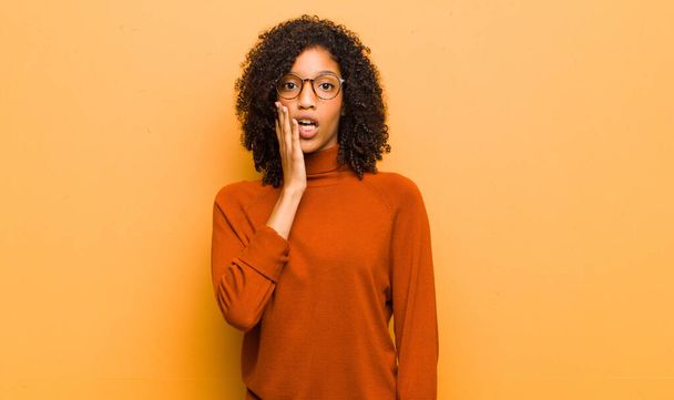 young pretty black woman feeling shocked and astonished holding face to hand in disbelief with mouth wide open against orange wall - Zdjęcie, obraz