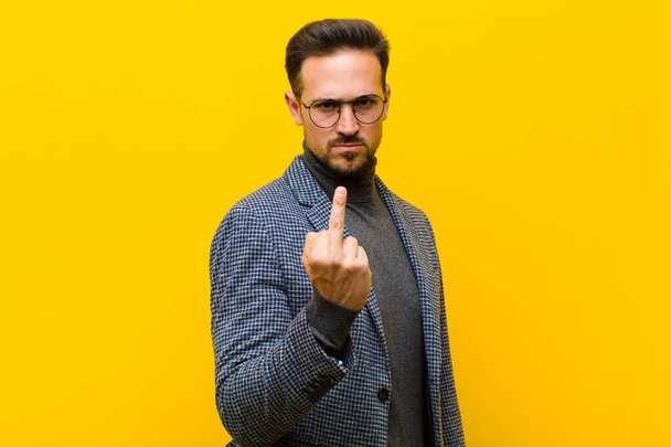 young handsome man feeling angry, annoyed, rebellious and aggressive, flipping the middle finger, fighting back against orange wall - Foto, Bild