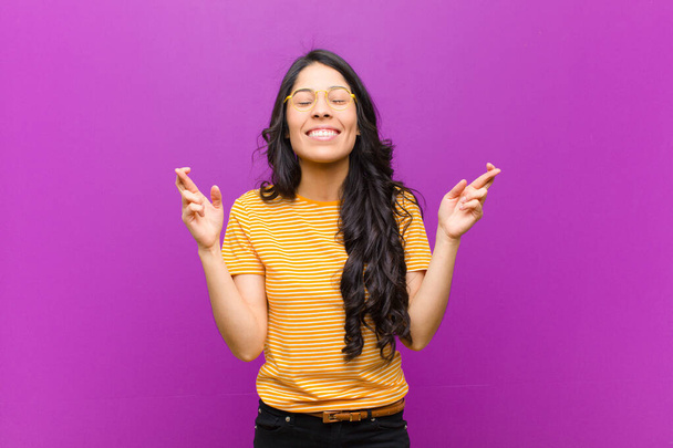 young pretty latin woman feeling nervous and hopeful, crossing fingers, praying and hoping for good luck against purple wall - Fotoğraf, Görsel