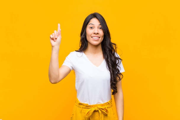 mladá hezká latinská žena se vesele a šťastně usmívá a ukazuje jednou rukou nahoru, aby kopírovala prostor na oranžové stěně - Fotografie, Obrázek