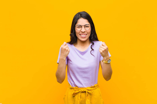 young pretty latin woman shouting triumphantly, laughing and feeling happy and excited while celebrating success against orange wall - Photo, image