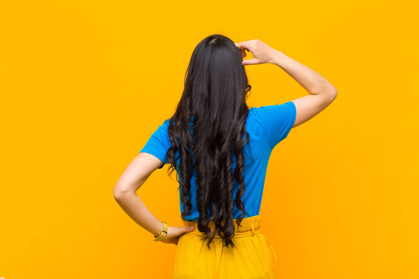 young pretty latin woman feeling clueless and confused, thinking a solution, with hand on hip and other on head, rear view against orange wall - Fotoğraf, Görsel
