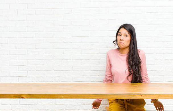 young  pretty latin woman feeling clueless and confused, having no idea, absolutely puzzled with a dumb or foolish look sitting in front of a table - 写真・画像