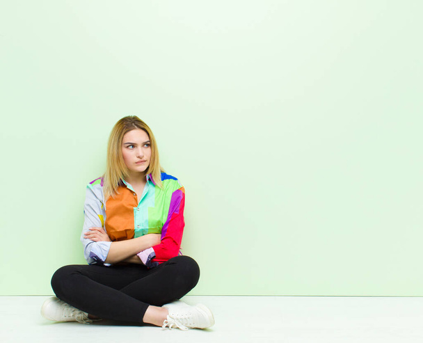 young blonde woman doubting or thinking, biting lip and feeling insecure and nervous, looking to copy space on the side sitting on the floor - Foto, Imagen