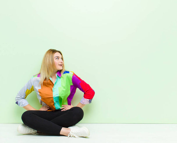 young blonde woman looking happy, cheerful and confident, smiling proudly and looking to side with both hands on hips sitting on the floor - 写真・画像