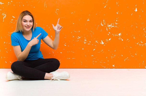 young blonde woman feeling joyful and surprised, smiling with a shocked expression and pointing to the side sitting on the floor - Photo, image