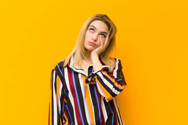 young blonde pretty woman feeling bored, frustrated and sleepy after a tiresome, dull and tedious task, holding face with hand against orange wall - 写真・画像