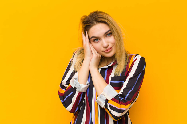 young blonde pretty woman feeling in love and looking cute, adorable and happy, smiling romantically with hands next to face against orange wall - Photo, image