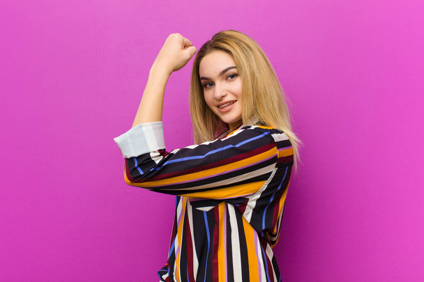 young blonde woman feeling happy, satisfied and powerful, flexing fit and muscular biceps, looking strong after the gym against purple wall - Фото, изображение