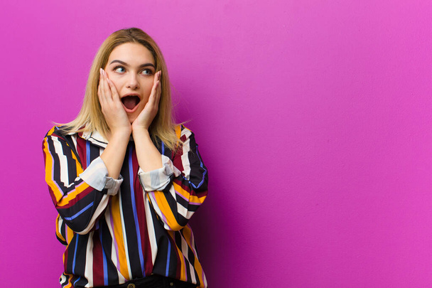 young blonde woman feeling happy, excited and surprised, looking to the side with both hands on face against purple wall - Foto, Imagem