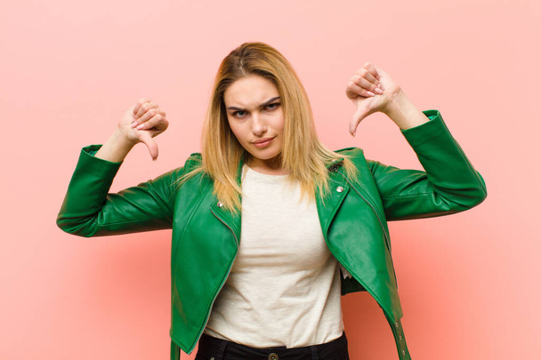 young pretty blonde woman looking sad, disappointed or angry, showing thumbs down in disagreement, feeling frustrated against pink flat wall - Foto, imagen
