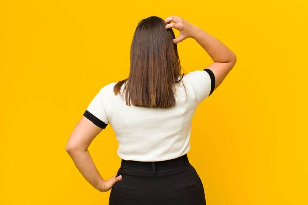 young pretty woman feeling clueless and confused, thinking a solution, with hand on hip and other on head, rear view isolated against orange wall - Photo, image