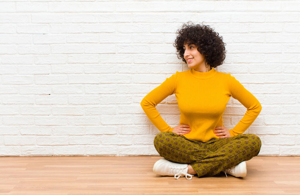young pretty afro woman looking happy, cheerful and confident, smiling proudly and looking to side with both hands on hips sitting on the floor - Photo, Image