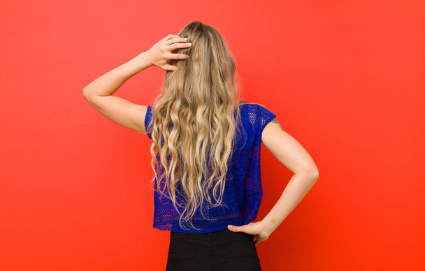 young blonde woman feeling clueless and confused, thinking a solution, with hand on hip and other on head, rear view against red wall - Photo, Image